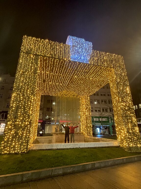 Two children pose beneath an enormous walk through Christmas present covered in thousands of golden lights. A glowing blue bow tops the festive Luxembourg holiday display.