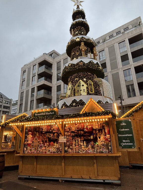A wooden Luxembourg Christmas market stall filled with ornaments and holiday decorations stands beneath a towering illuminated Christmas pyramid. Signs on the stall read "Beautiful Market" and "Gifts Cadeaux Decorations Merry Christmas".