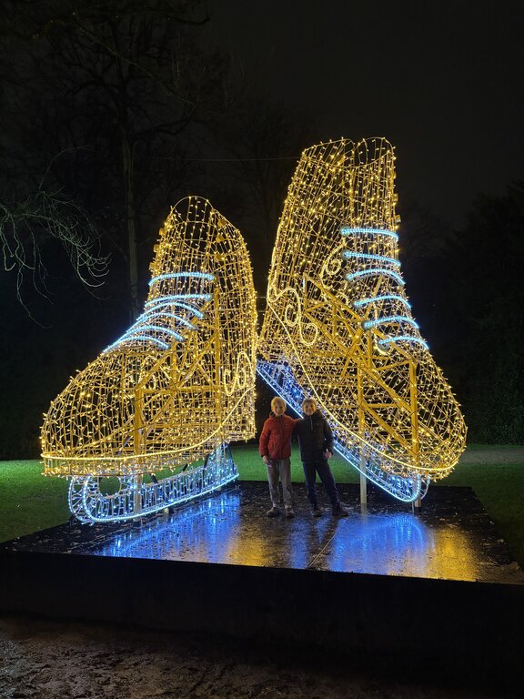 Two children stand between enormous ice skate sculptures made from golden and blue Christmas lights in Luxembourg. The oversized illuminated skates tower over them against the dark night sky.