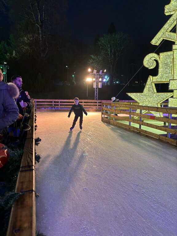 A child skates across an outdoor ice rink in Luxembourg with colorful spotlights and a giant illuminated Christmas decoration nearby. Spectators watch from behind the wooden railing.