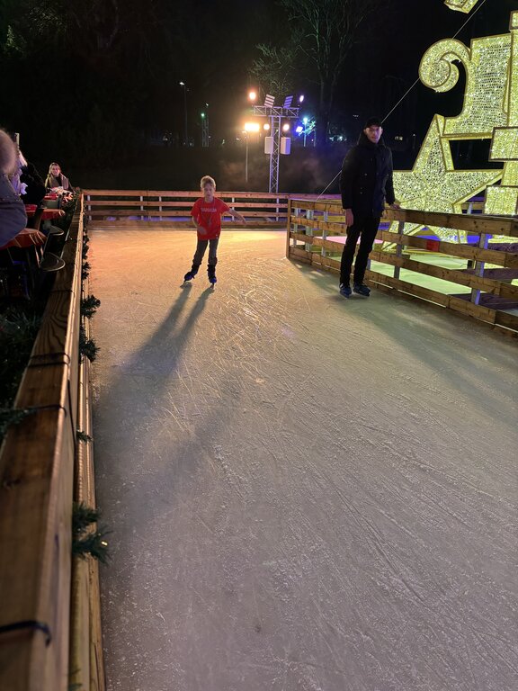 A child in a red shirt skates along a festive outdoor ice rink in Luxembourg. Warm spotlights and an oversized golden Christmas decoration illuminate the rink at night.
