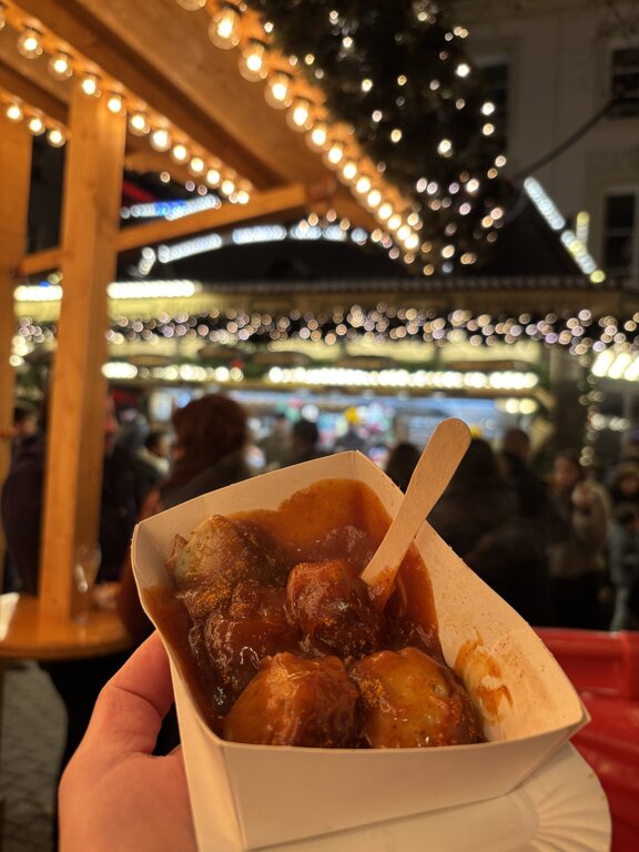 A serving of sliced sausage covered in rich brown curry sauce and held in front of a warmly lit Luxembourg Christmas market stall. Strings of glowing lights and market visitors create a festive background.