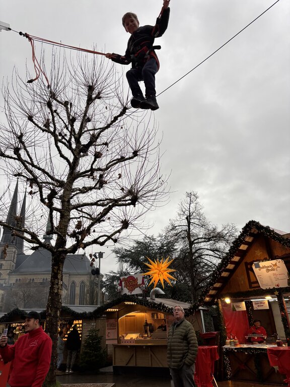 A child rides high above a Luxembourg Christmas market on a bungee trampoline attraction. Wooden market stalls and a glowing Christmas star sit below beneath a cloudy winter sky.