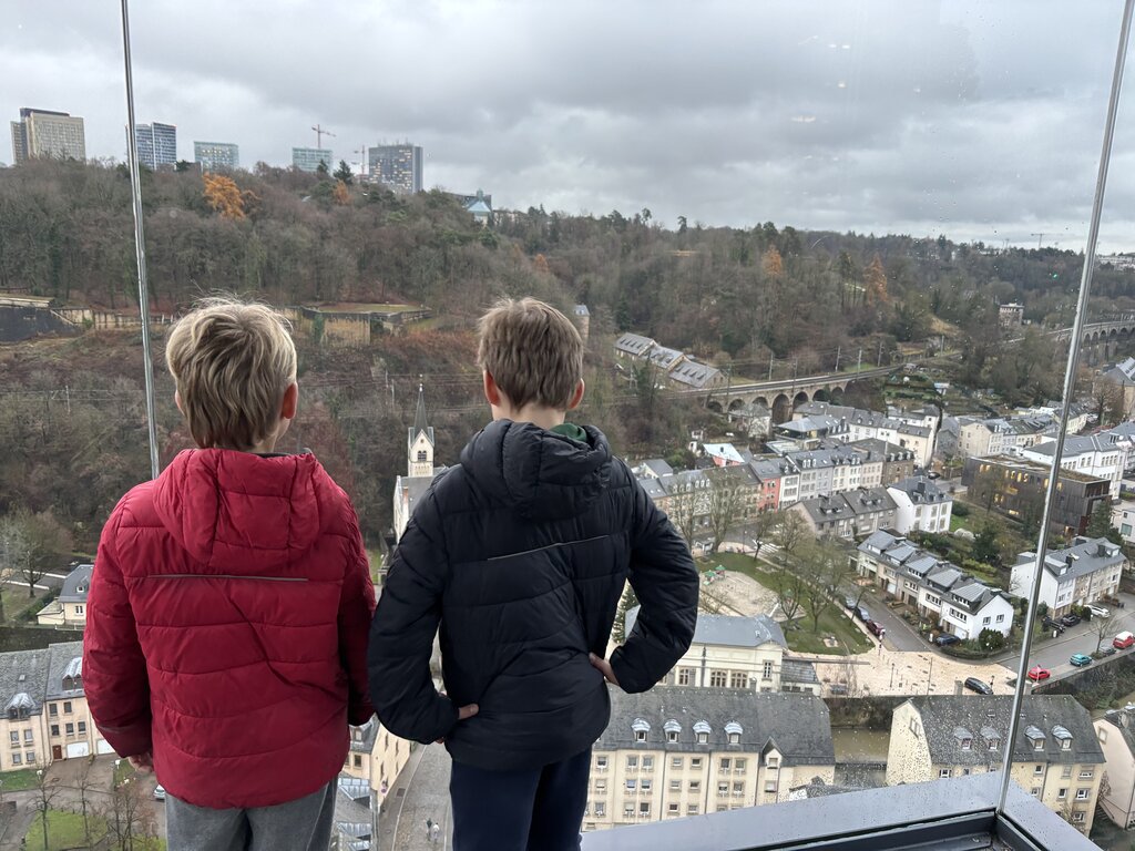 Two children look through a large glass window of the elevator over the lower neighbourhoods of Luxembourg City. Historic buildings and a stone viaduct spread across the valley below with wooded hills in the distance.