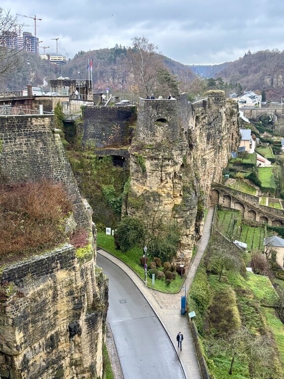Dramatic rocky cliffs and historic stone fortifications rise above a winding road in Luxembourg City. Walking paths and gardens weave along the base of the cliffs with wooded hills beyond.